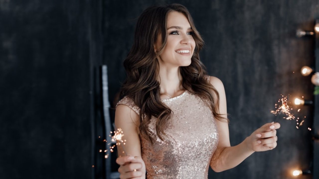 Girl with a sparkler near the Christmas tree. A cheerful young woman with a cute smile in a beige dress is standing and holding a sparkling sparkler in the hands against the background of the Christmas tree. Beautiful girl celebrates Christmas.